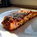Close-up of a flaky, seasoned air fryer salmon fillet resting on a white plate, showing its moist interior.