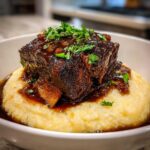 A close-up of a tender braised short ribs piece covered in rich sauce, served atop creamy polenta and garnished with fresh parsley.