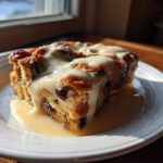 A decadent slice of bread pudding topped with a thick, creamy sauce, served on a white plate.