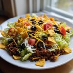 A vibrant, fully loaded taco salad featuring seasoned ground beef, lettuce, tomatoes, black olives, cheese, and tortilla chips.