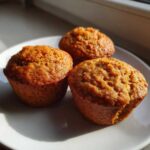 Three freshly baked banana protein muffins with a golden-brown, textured top sitting on a white plate near a window.