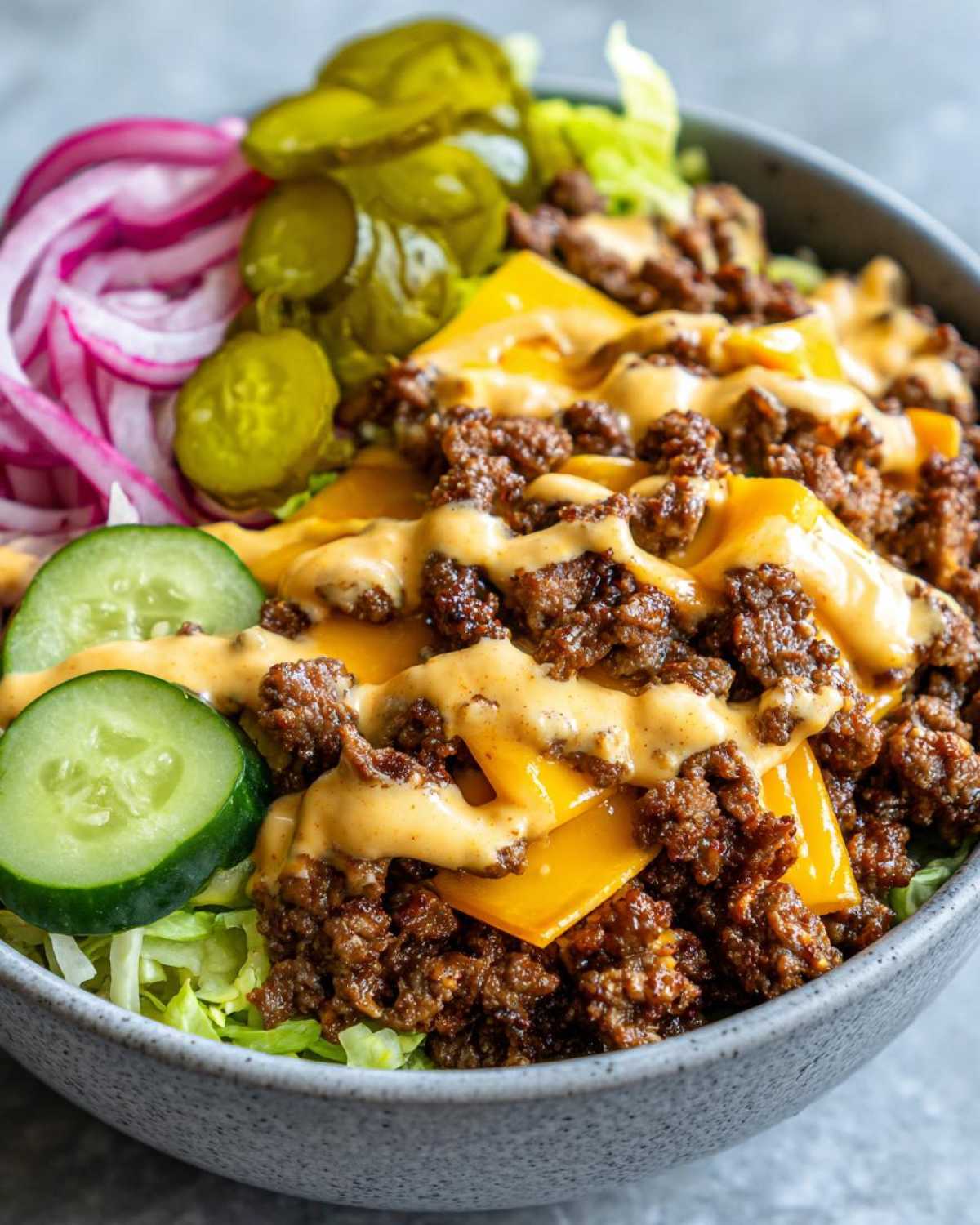 A close-up of a delicious cheeseburger bowl featuring seasoned ground beef, melted cheese, and a drizzle of sauce over lettuce, with pickles and red onion on the side.