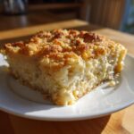 A close-up of a square slice of cheesy broccoli casserole with a golden brown crumb topping on a white plate.