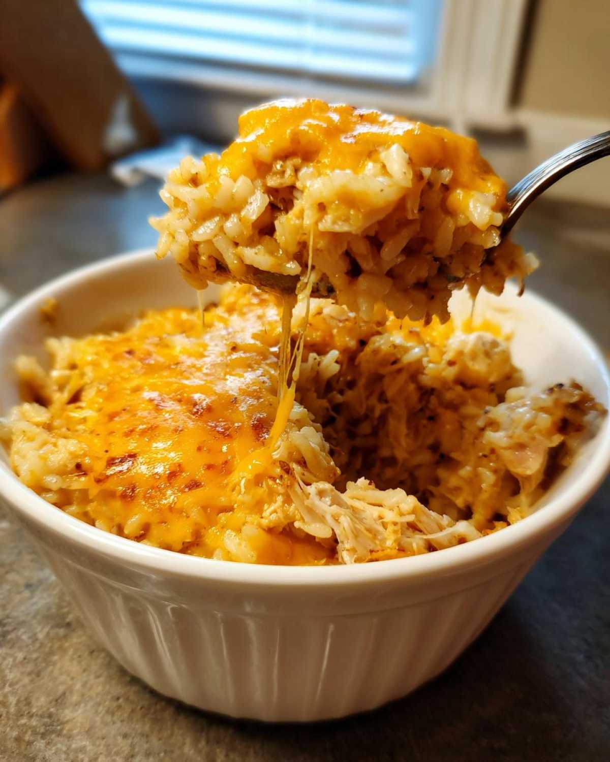 A spoonful of cheesy chicken and rice lifted from a white bowl, showing melted cheese strings.