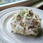 A scoop of creamy cottage cheese chicken salad mixed with red onion and topped with fresh dill on a white plate.