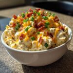 A white bowl filled with creamy million dollar dip, topped generously with shredded cheddar cheese, bacon bits, and green onions.