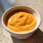 Close-up of smooth, bright orange nacho cheese sauce filling a small white ramekin, sitting on a light countertop.