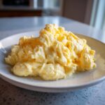 A close-up of a generous serving of soft, creamy scrambled eggs piled high on a light-colored plate.