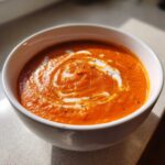 A close-up of a vibrant orange bowl of creamy tomato bisque, garnished with a swirl of white cream and pepper.