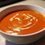 A close-up of a bowl of vibrant orange tomato bisque, garnished with a swirl of cream.
