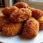 A pile of golden-brown, crispy baked Jalapeño poppers on a white plate, one is broken open showing the cheesy filling.