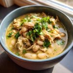 A close-up of a bowl of rich crockpot white chicken chili topped with shredded chicken, white beans, and fresh cilantro.