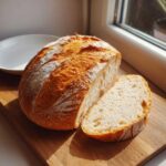 A freshly baked, crusty Italian bread loaf, dusted with flour, sitting on a wooden board with one slice cut.