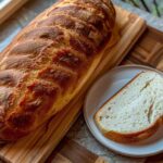 A freshly baked, crusty Italian bread loaf resting on a wooden cutting board next to a single slice on a white plate.