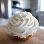 A single cupcake topped with a tall, perfectly piped swirl of white cream cheese frosting, sitting on a wooden board.
