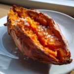 Close-up of a perfectly baked sweet potato, split open to show the fluffy orange flesh topped with coarse sea salt.