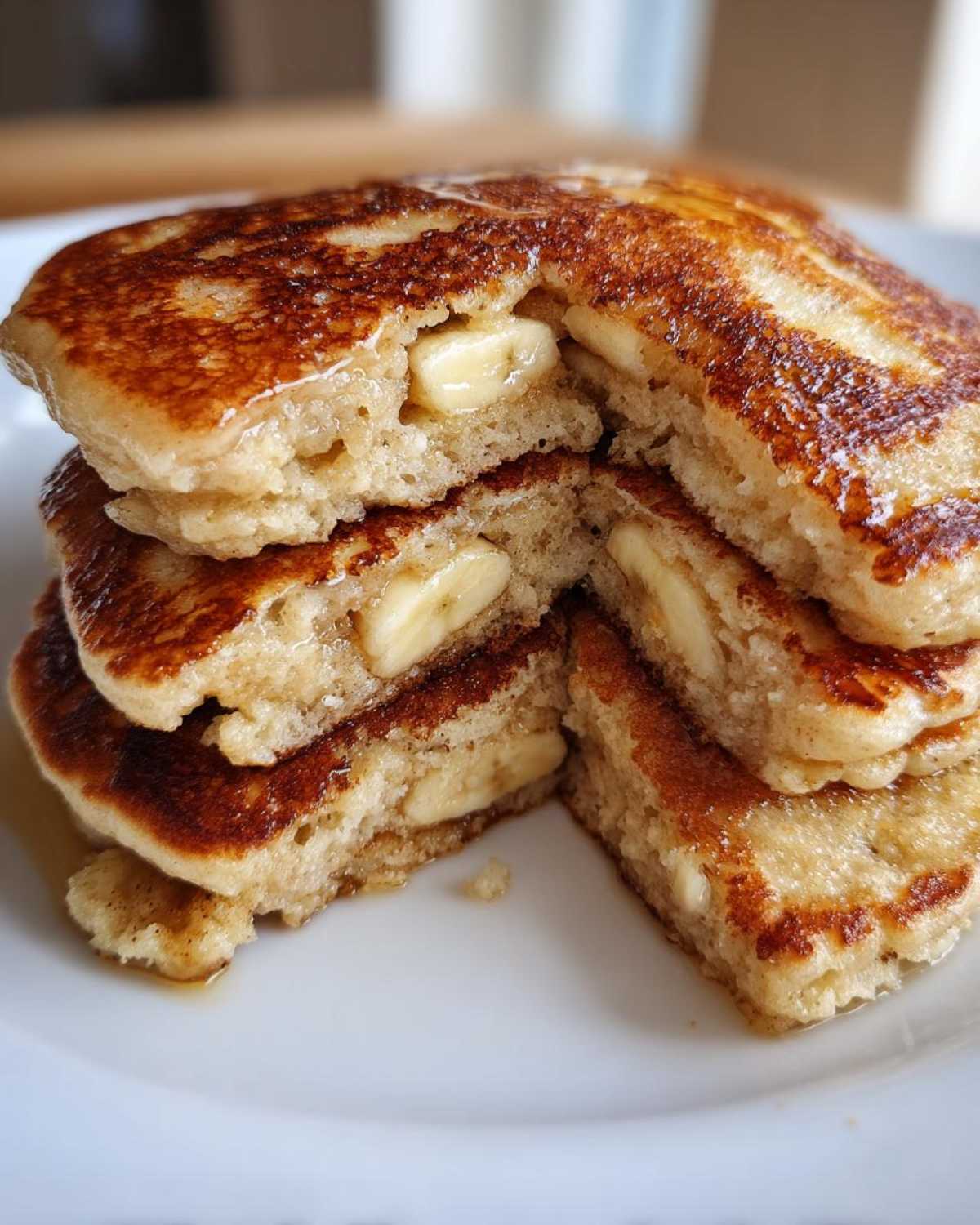 A stack of fluffy banana pancakes cut open to show slices of banana baked inside, drizzled with syrup.