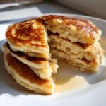 Close-up of a stack of fluffy banana pancakes cut open to show the airy interior, drizzled with syrup.