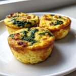 Three golden brown, fluffy egg bites topped with spinach and bacon pieces served on a white plate.