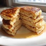 A stack of fluffy greek yogurt pancakes with a piece cut out, drizzled with syrup.