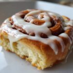 Close-up of a fluffy, golden-brown cinnamon roll generously topped with white icing, with a small bite missing.