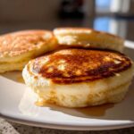 Close-up of three incredibly fluffy pancakes with golden-brown tops, drizzled with syrup on a white plate.