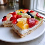 Close-up of a slice of fruit pizza sugar cookie topped with cream cheese frosting and fresh fruit.