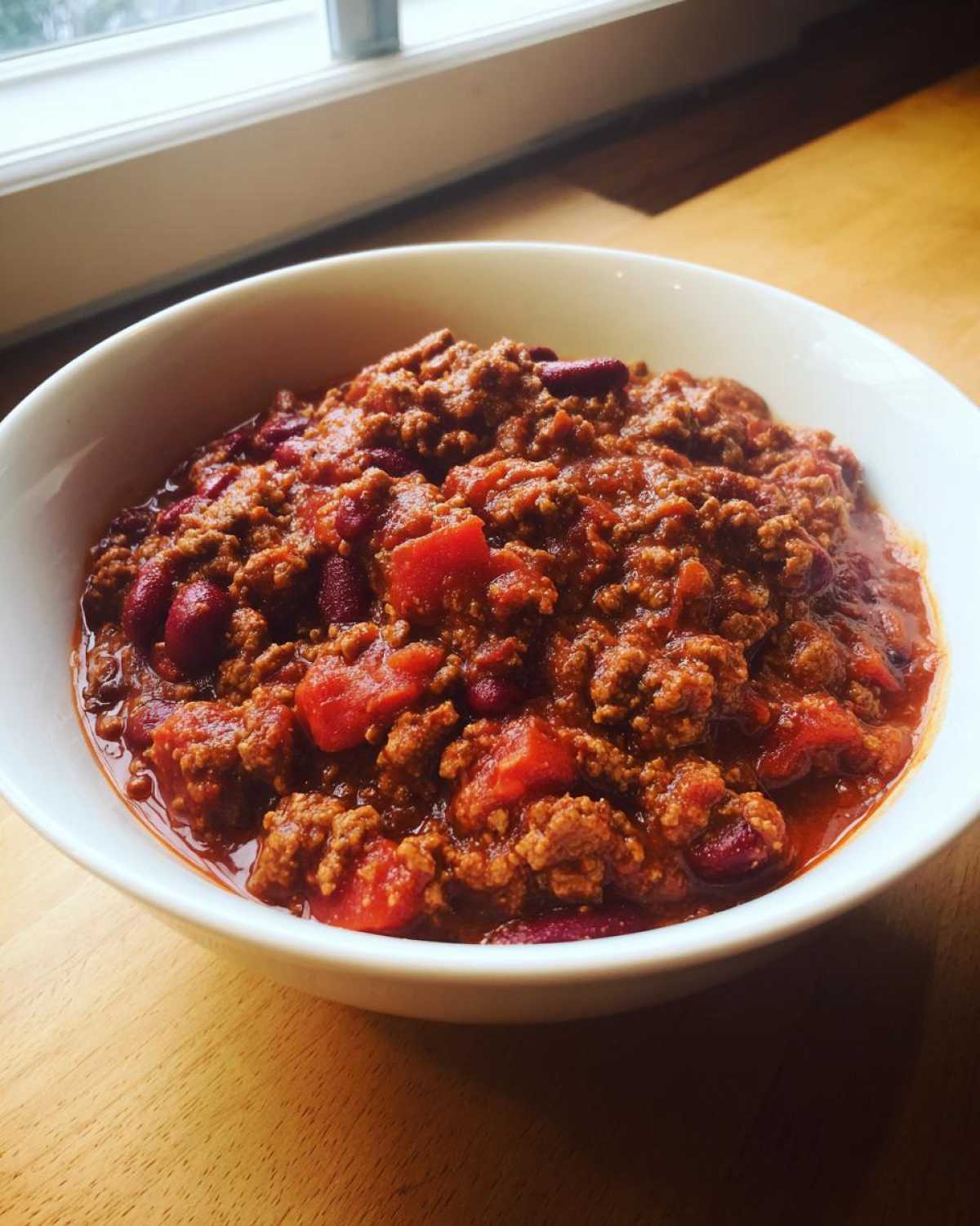 A close-up of a white bowl filled with rich, thick crockpot chili featuring ground meat, kidney beans, and tomatoes.