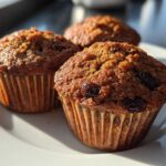 Close-up of three moist, dark honey bran muffins with visible raisins, sitting on a white plate in natural light.