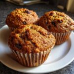 Close-up of three freshly baked honey bran muffins studded with raisins sitting on a white plate.