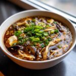 A close-up bowl of rich, dark hot and sour soup recipe, garnished heavily with fresh green onions.