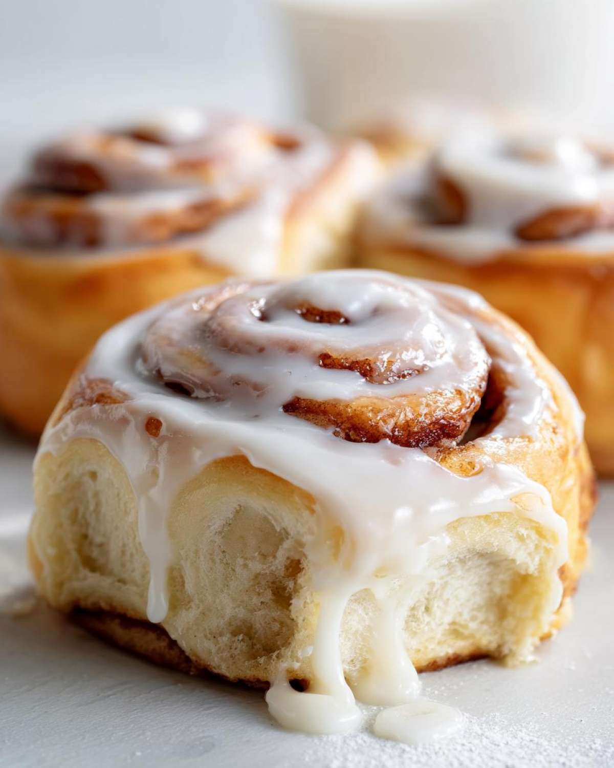 A close-up of a soft, freshly baked cinnamon roll covered in thick white icing dripping down the side.