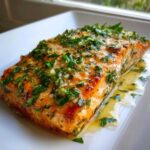 A close-up of a perfectly cooked baked salmon recipe fillet topped with a bright green garlic and herb butter sauce.
