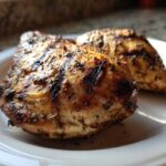 Two perfectly seared and seasoned greek chicken breasts resting on a white plate.