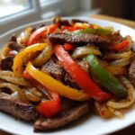 Close-up of juicy steak fajitas featuring strips of seasoned steak mixed with colorful sautéed bell peppers and onions on a white plate.