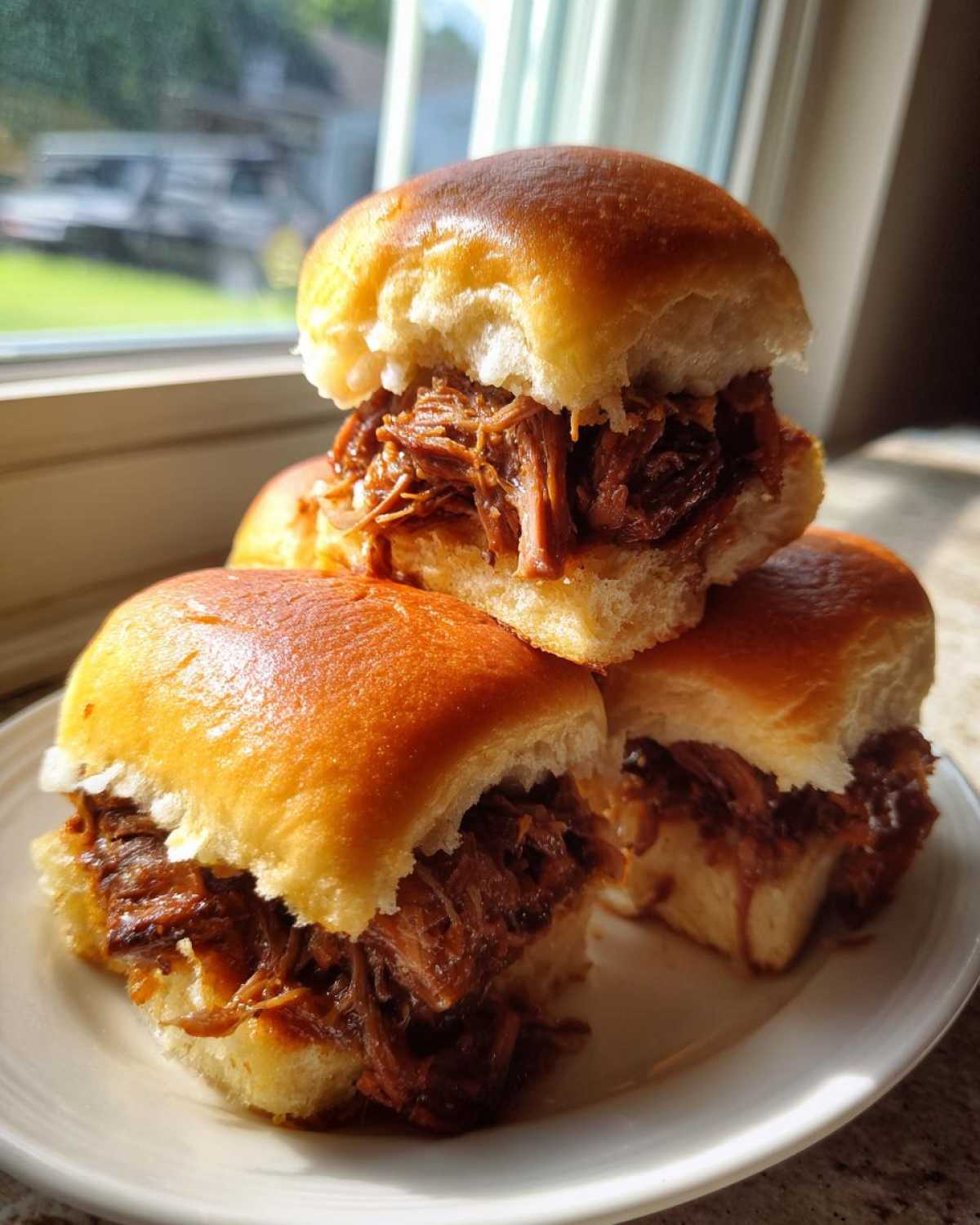 Three delicious mississippi pot roast sliders stacked on a white plate, filled with shredded, saucy beef.