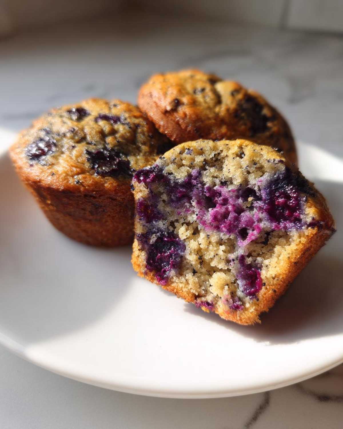Close-up of three blueberry protein muffins, one cut in half showing the moist interior and bright purple blueberries.