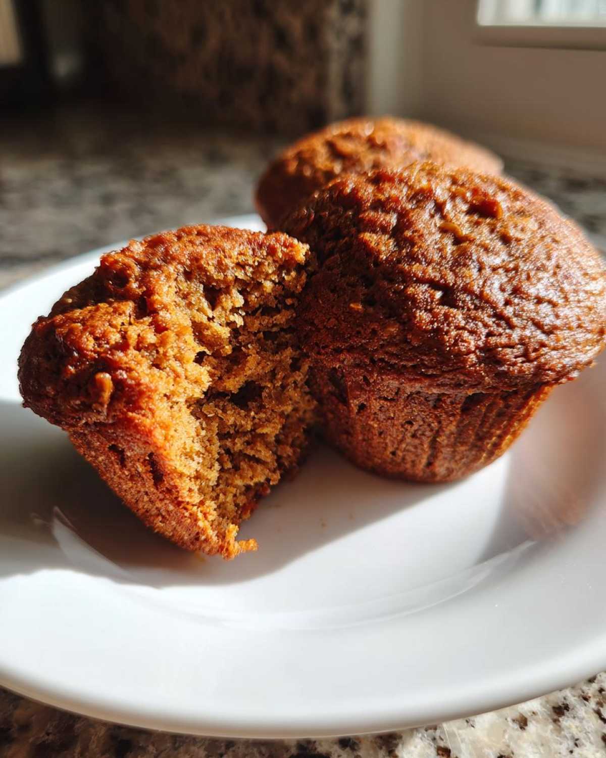 Close-up of moist bran muffins on a white plate, one muffin is broken open showing the texture.