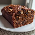 A close-up of a thick slice of moist date nut bread, studded with dates and topped with walnuts, served on a white plate.