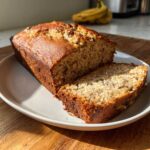 A loaf of moist sugar free banana bread with one slice cut and resting against the main loaf on a white plate.