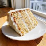 A close-up slice of moist vanilla cake recipe with white frosting, sitting on a white plate in bright sunlight.