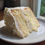 A close-up slice of moist vanilla cake recipe with thick white frosting, sitting on a white plate near a sunny window.