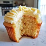 Close-up of a vanilla cupcakes with a bite taken out, showing the fluffy, moist crumb and thick vanilla frosting.