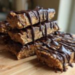 A stack of homemade, no-bake protein bars drizzled generously with melted dark chocolate, resting on a wooden cutting board.