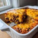 A square serving of cowboy cornbread casserole being lifted from a white baking dish, showing layers of meat, corn, and melted cheese.