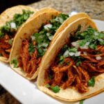 Close-up of three soft corn tortillas filled with shredded chicken tacos, topped with fresh cilantro and diced white onion.