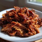 A mound of saucy, shredded Slow Cooker Pulled Pork piled high on a white plate, catching bright sunlight.
