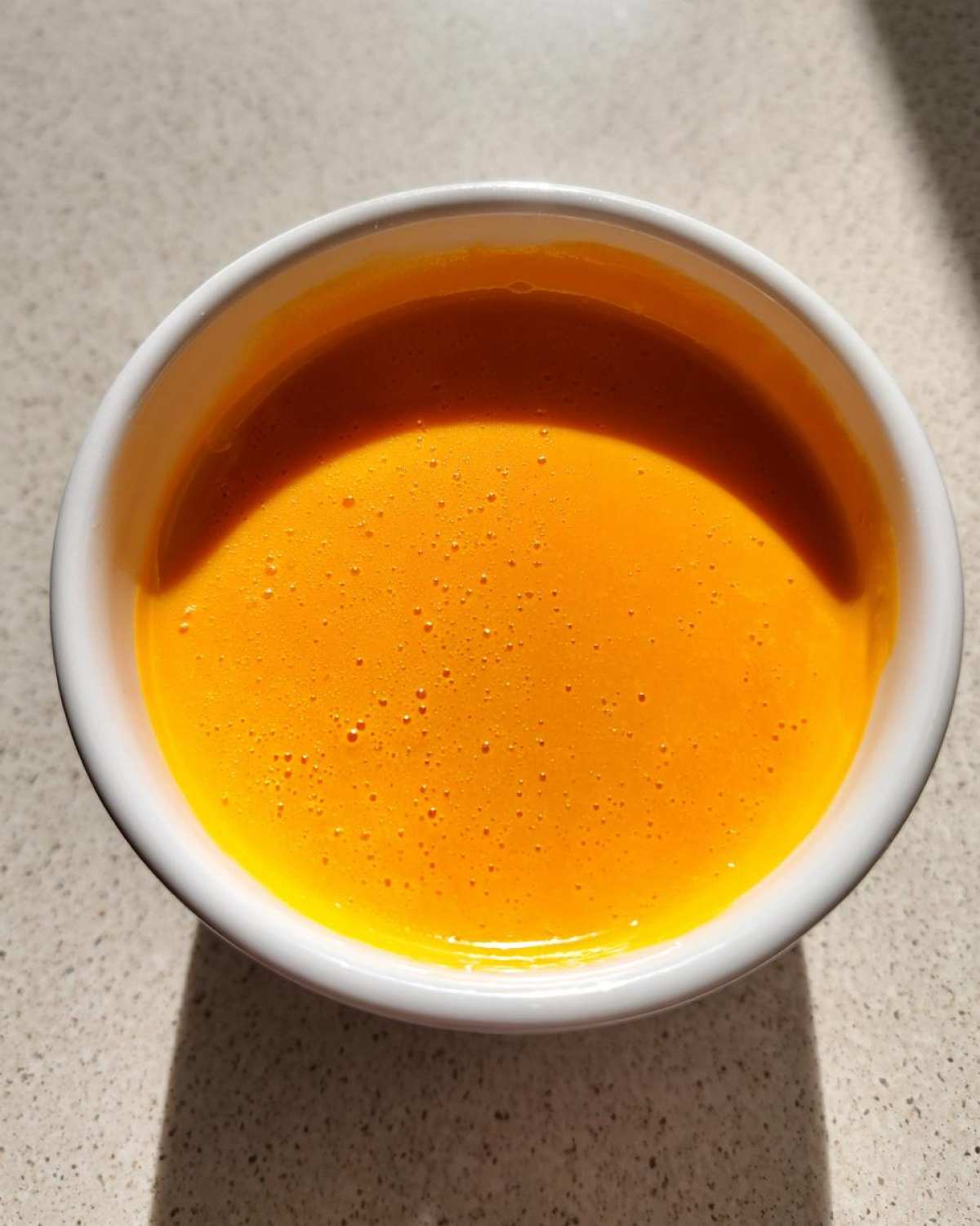 Overhead view of vibrant orange, smooth nacho cheese sauce in a small white bowl, glistening under sunlight.