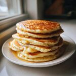 A tall stack of five golden brown, fluffy pancakes drizzled generously with maple syrup on a white plate.