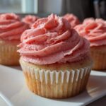 A close-up of one perfectly frosted strawberry cupcake with bright pink strawberry frosting, with more strawberry cupcakes blurred in the background.